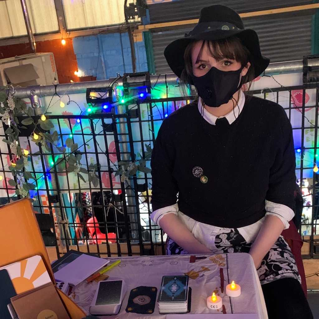 Imogen sits at a small table with some tarot cards, they are wearing a hat and a respirator.